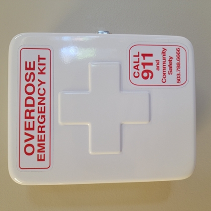 White naloxone emergency kit case with red lettering affixed to wall.